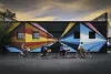 Bicyclists ride in front of a mural at sunset.
