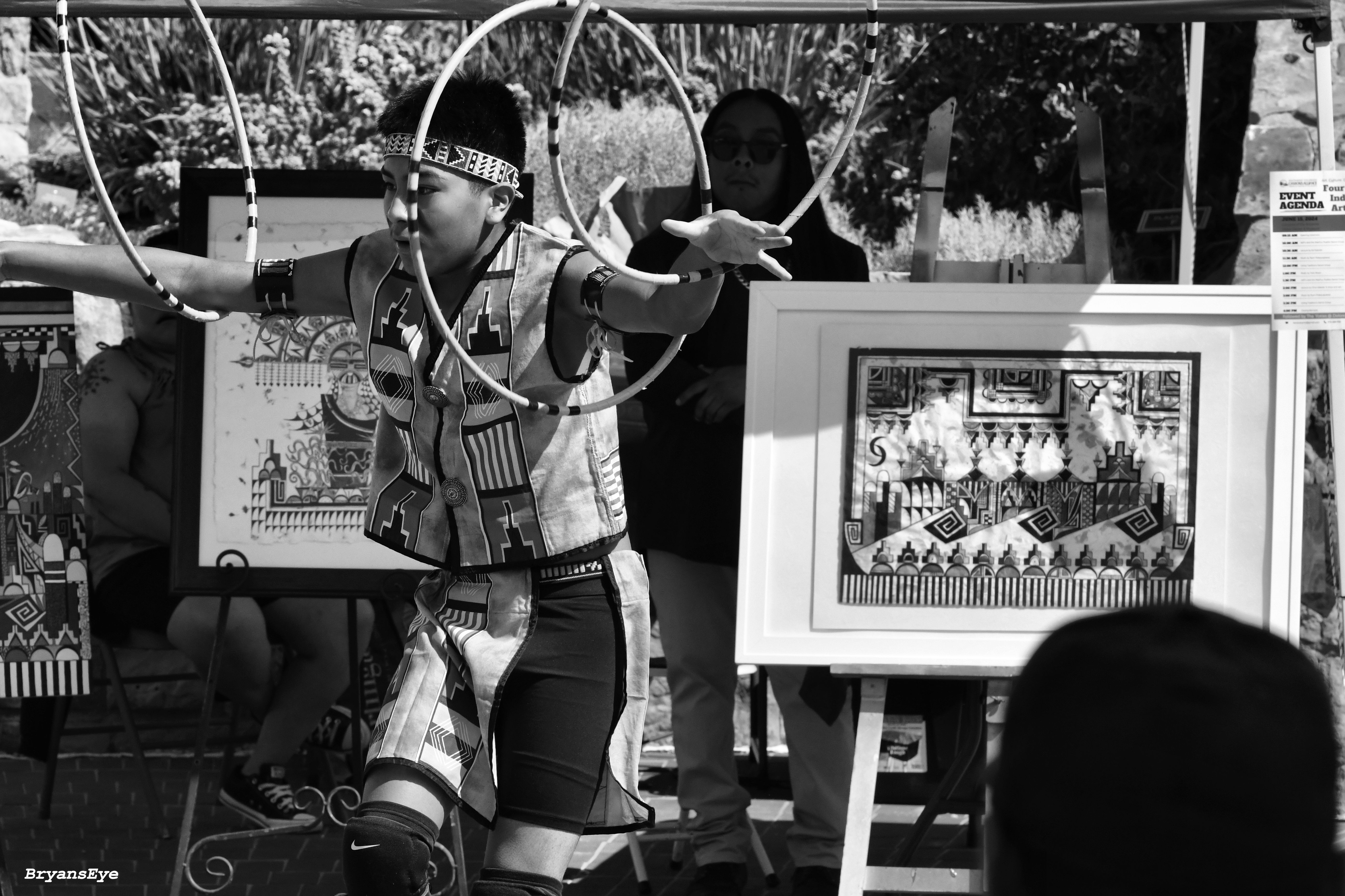 Black and White image of a child dancing in traditional native attire. In front of a community marketplace
