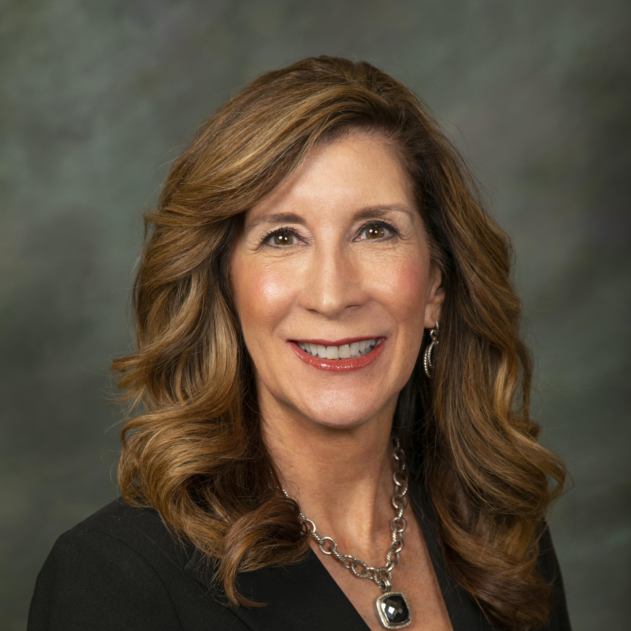 Woman with long brown hair, black shirt and suit jacket with a silver nacklace