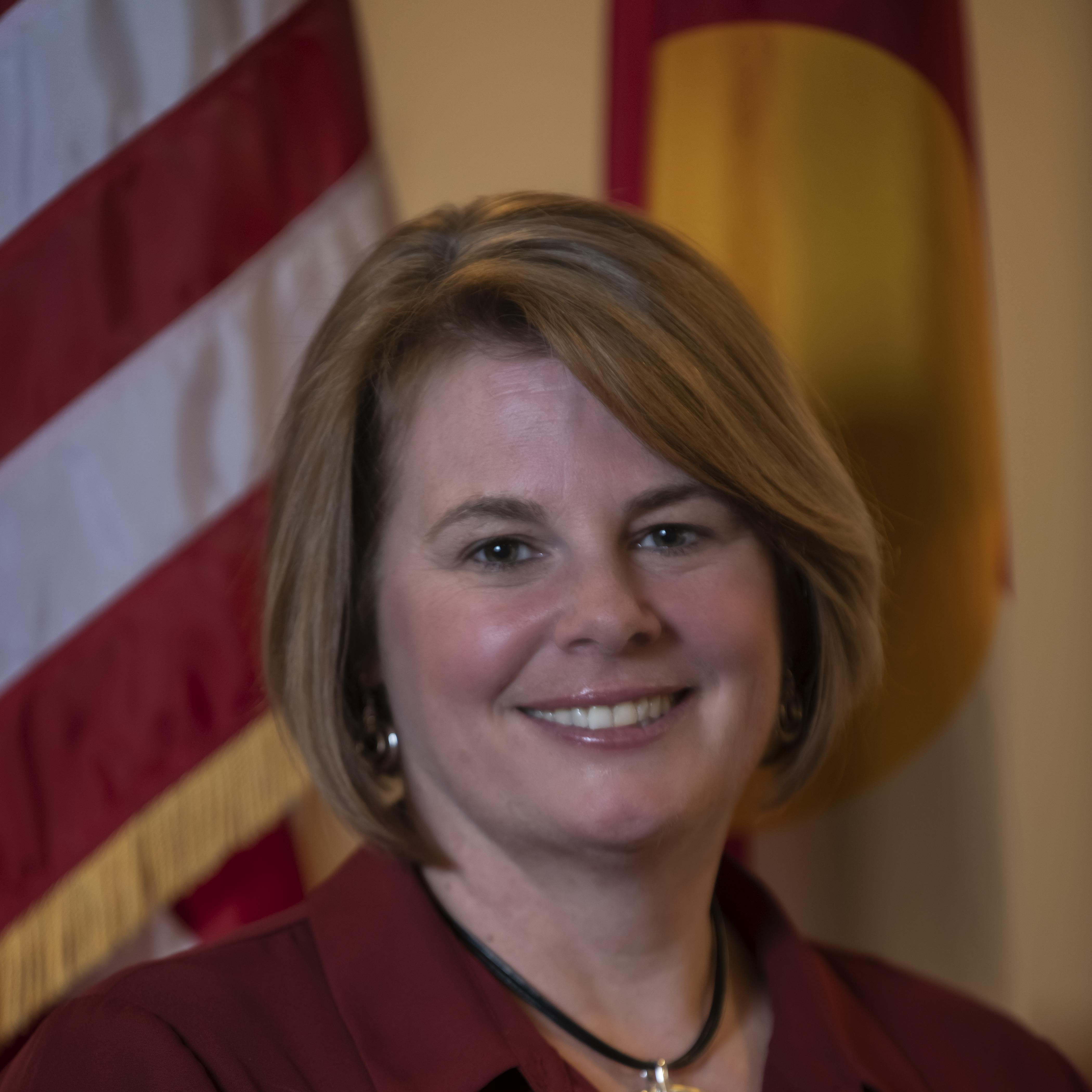 Woman smiling and wearing a dark red button down shirt with a black necklace and gold piece of jewelry hanging from the black necklace. She has light brown hair and is standing in front of a U.S. flag and the Colorado Flag. 