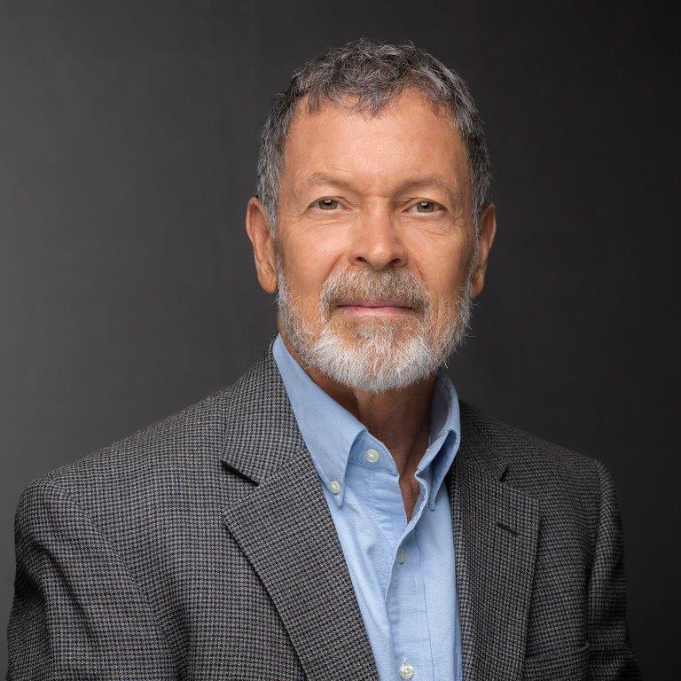 Man wearing a blue button down, checkered grey suit jacket, black and gray short hair cut and a gray beard with a slight smile.