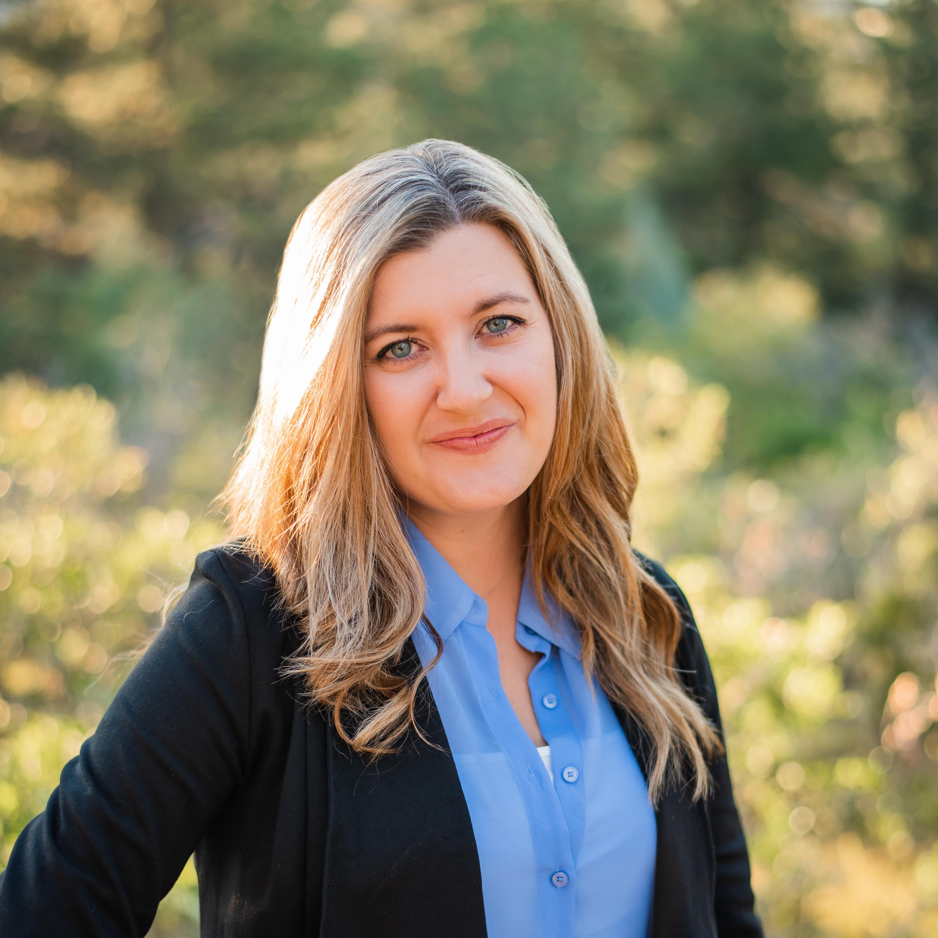 Phot of Ashley Mount smiling and wearing a black business suit with a blue collard shirt. She has one had on her hip and the background is green/yellow greenery. 