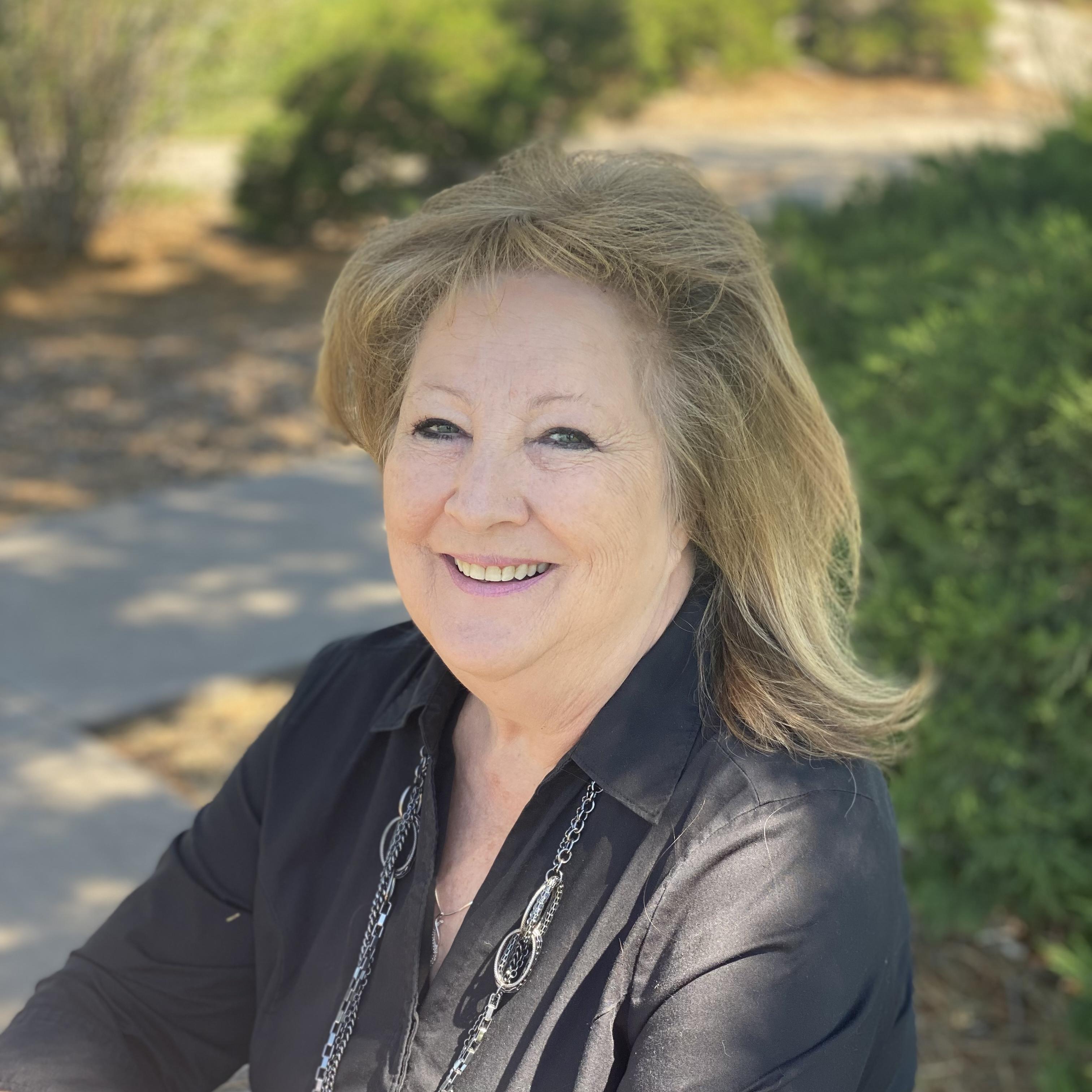 Image of Dena Daniel siting in a park bench. She has a black shirt, blonde hair, and a necklace