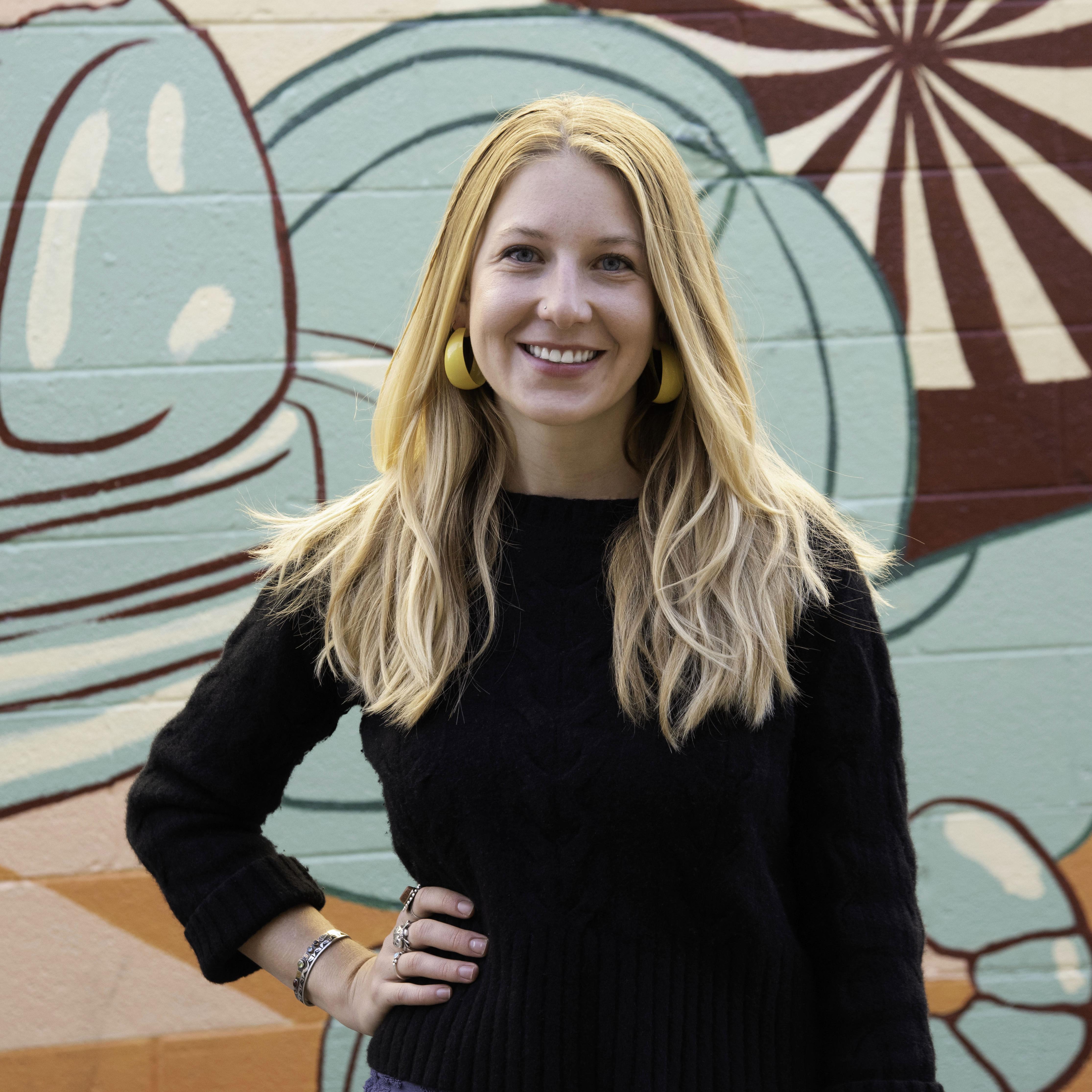 Image of Emma in front of an abstract mural. She has a black shirt and blue pants/skirts. She has blonde hair and yellow earrings. Her right hand is on her hip. 