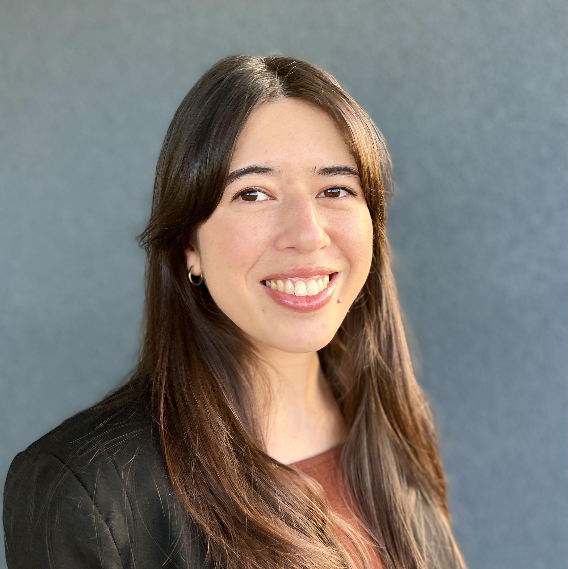 Headshot image of Michelle. She has a dark business jacket with a brown top. She has dark brown hair and is wearing small hoop earrings. She is smiling and showing her teeth. 