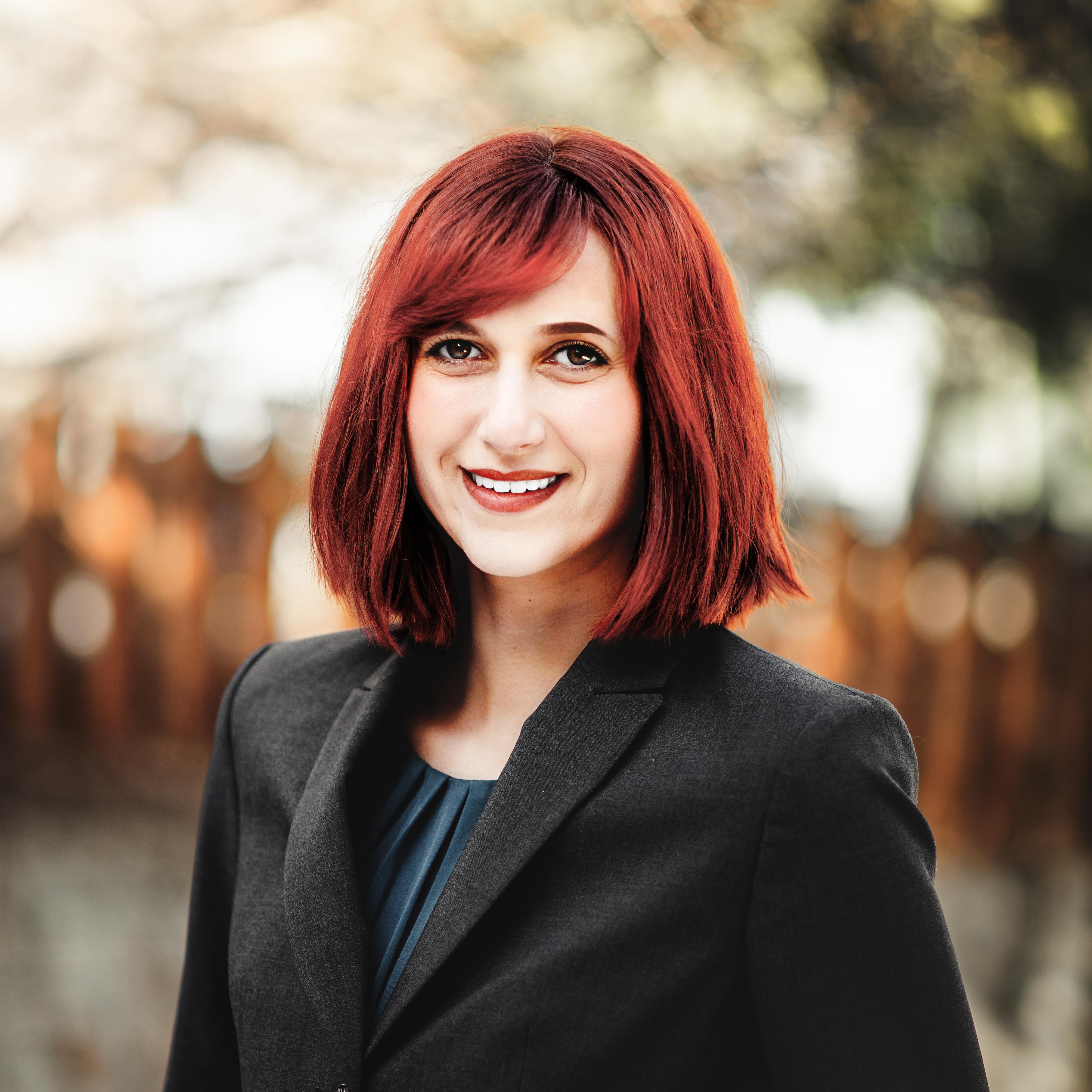 Image of Arielle Brachfeld. She has a black suit, red hair, and is smiling. The background image is blurred of an outdoor setting with a brown fence. 