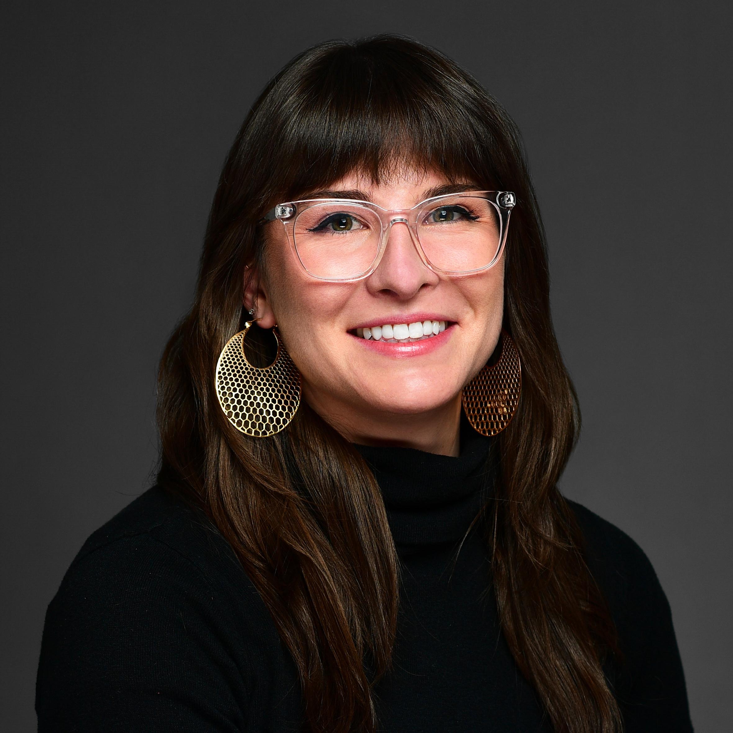 Women smiling, long brown hair, wearing a black turtle neck and clear framed glasses