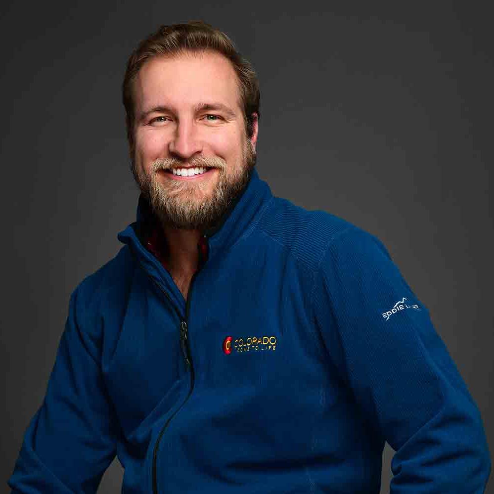 Image of Conor Hall smiling and wearing a blue jacket. She has blue eyes, dark blonde hair, a beard, and a mustache.