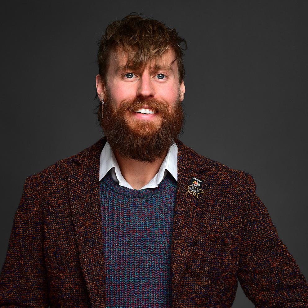 Headshot of Matthew Licina. He is wearing a brown business jacket, a blue sweater, and a white collard shirt. Matthew has brown hair, a beard-mustache combo, and a pin on is jacket.
