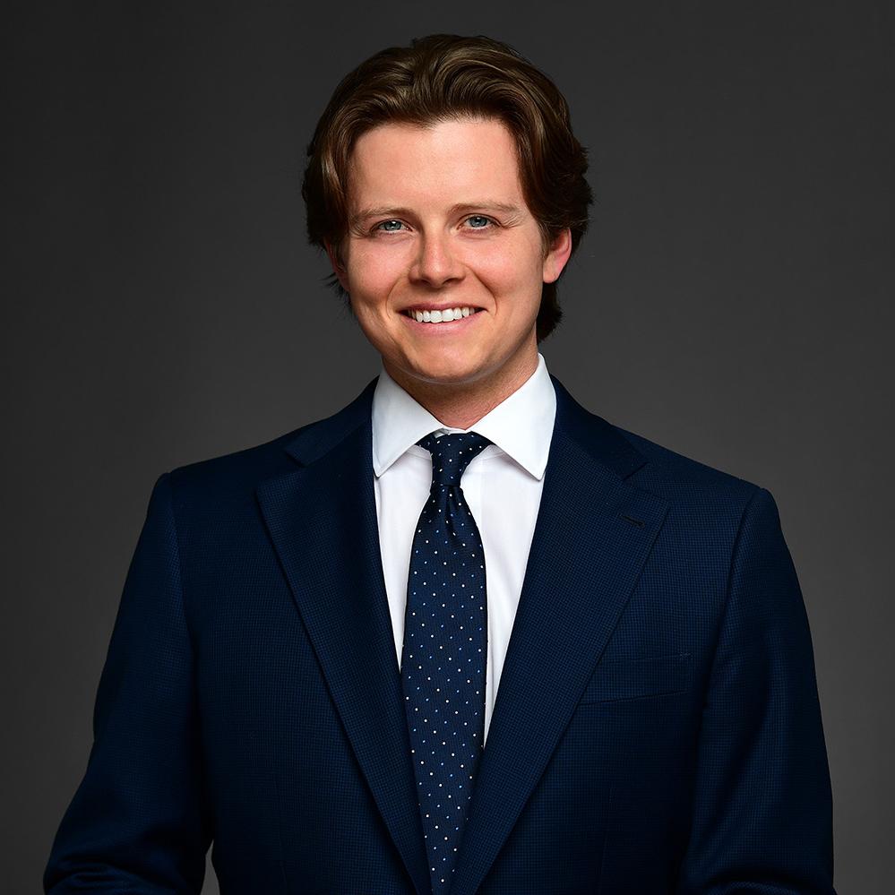 Headshot image of Xander Martin. He is wearing a dark blue business suit, blue tie with white dots, and white collard shirt. He is smiling and has brown hair. 