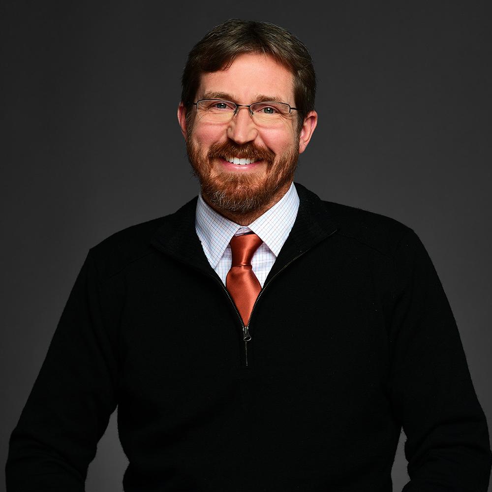 Eric Pynn is smiling and wearing a black jack with a white collard shirt and an orange tie. He is also wearing glasses and has a beard with a mustache. 