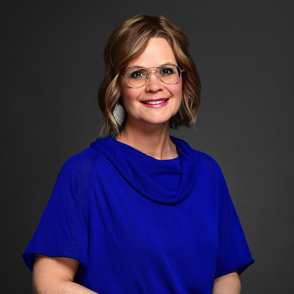 Jennifer Salvatore is smiling and wearing a blue blouse. She has on glasses, short blonde hair, and diamond shaped earrings. 