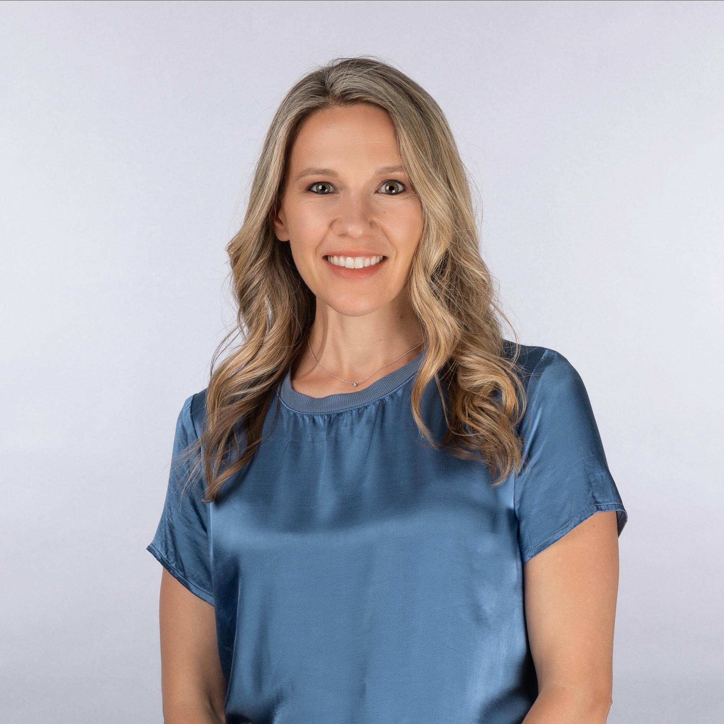 Head shot image of Allie Clark. She is wearing a blue velvet blouse and is smiling at the camera. Her backgroud is a white and silver color. She has blonde wavy hair that goes to the upper part of her arms. 