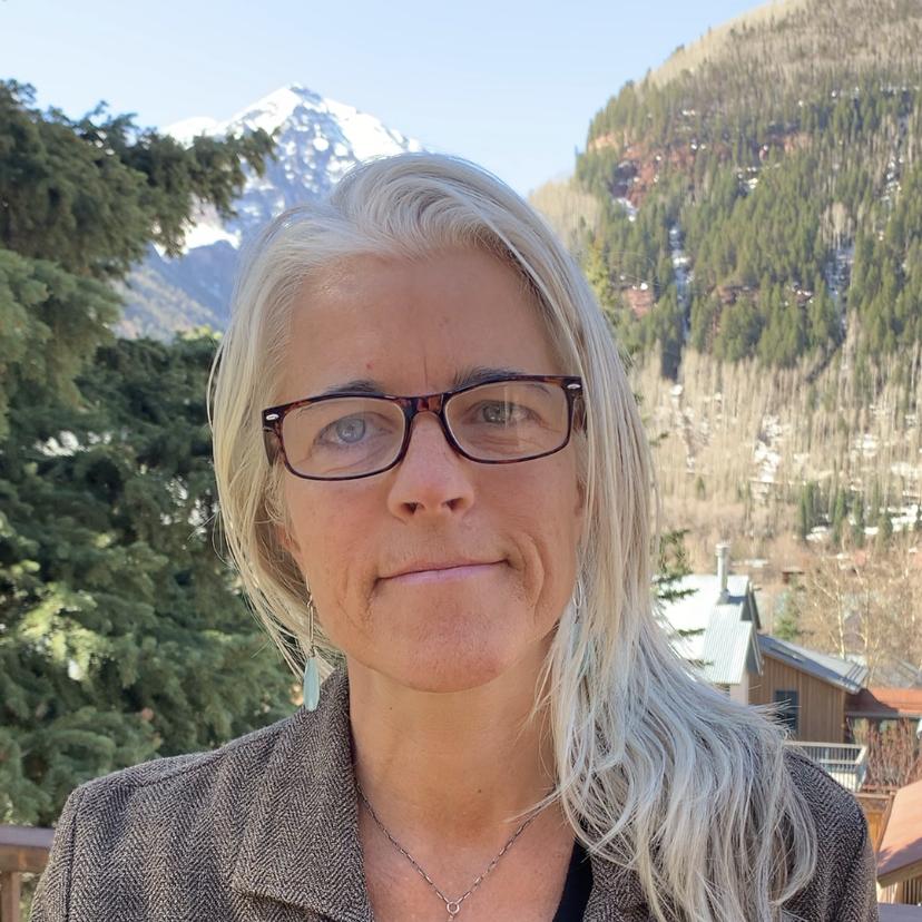 Hilary Cooper is smiling at the camera. She had brown glasses and light blonde hair that goes to her left shoulder. She is wearing a gray business jacket with a silver necklace. The background is a mountain, pine trees, and a log cabin. 