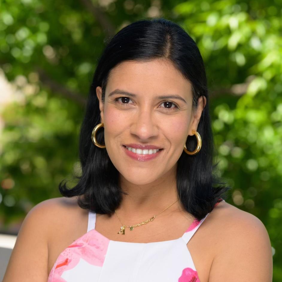 Andrea has medium short black hair and is wearing gold hoop earrings. She is smiling and has dark brown eyes. She has a necklace with the letter A on it and wearing a white tank top with roses on the tank top. The background is blurred trees. 