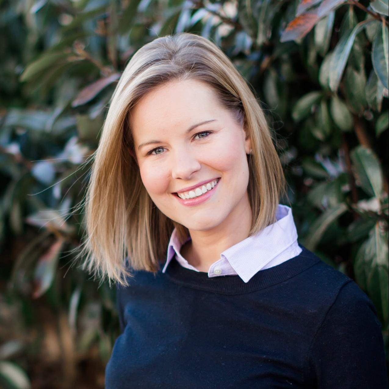 Marie has medium length blonde hair. She is tilting her head to view the camera and smiling. She has blue eyes and is wearing a white collard shirt. Over the white collard shirt she is wearing a dark navy blue sweater. The background is of greenery. 