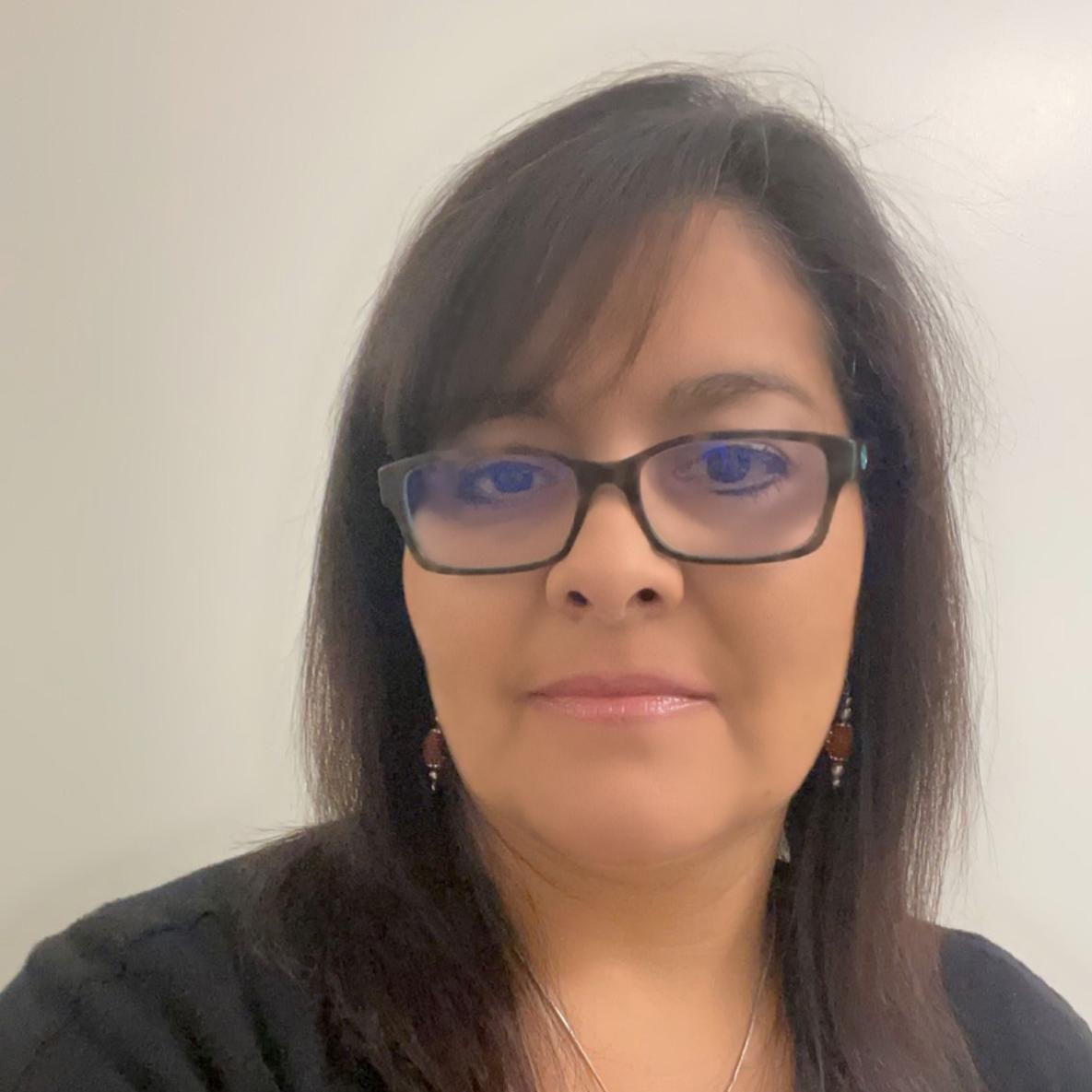 Stephanie Gonzales has medium length brown hair. She is wearing glasses, earings, a black cardigan, a necklace, and a textured blue shirt. They also have pink lip gloss and looking at the camera. 
