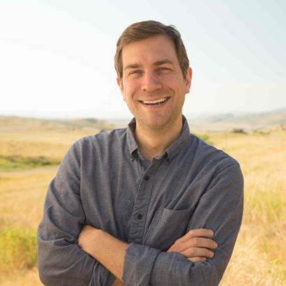 Jeff is crossing his arms and wearing a gray collard shirt. He has short brown hair and is smiling. HIs top teeth are showing and he recently shave his facial hair. The background is a plain field with some green hills.