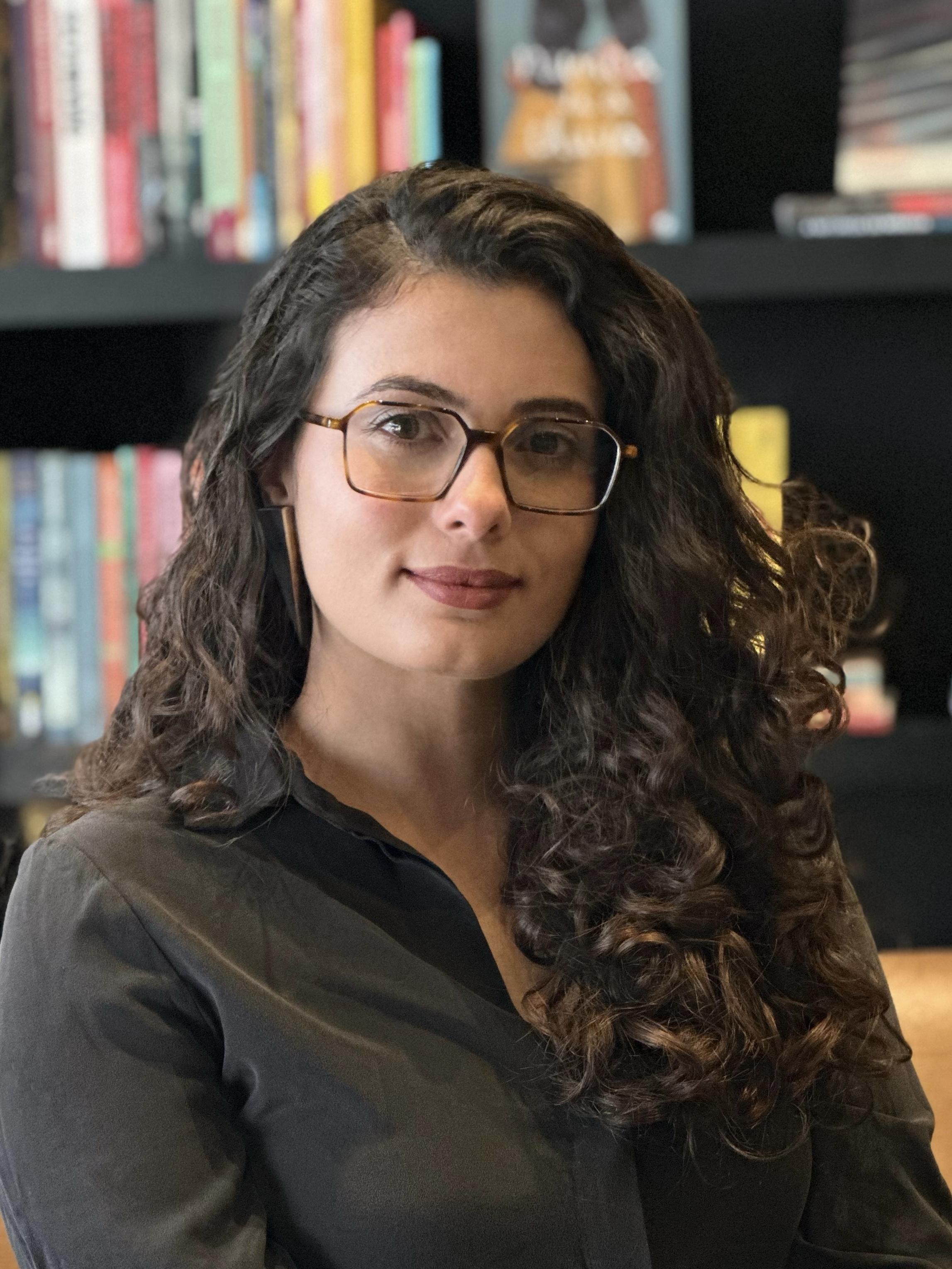 Júlia Martins Rodrigues wearing glasses and has curly hair while sitting in front of a bookshelf.