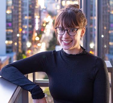 Emma Howard wearing a black turtleneck, standing in front of skyscrapers in early evening 