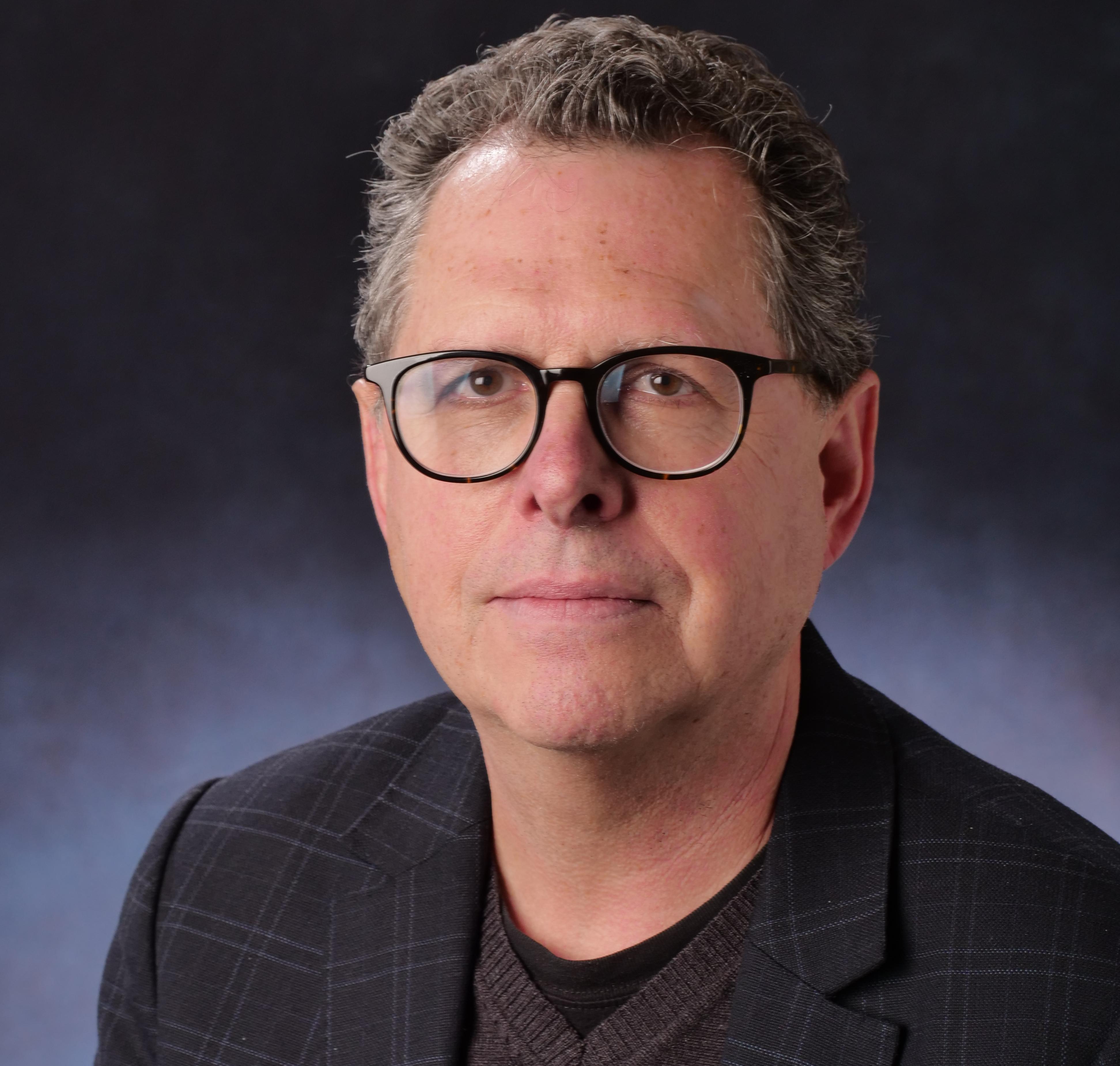 Man with grey hair and glasses, wearing a dark shirt and jacket on a dark background, looking into the camera.
