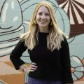 Image of Emma in front of an abstract mural. She has a black shirt and blue pants/skirts. She has blonde hair and yellow earrings. Her right hand is on her hip. 