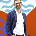 A white middle aged man with white shirt and blue suit jacket and glasses smiles in front of a red, white, and blue modern abstract mural