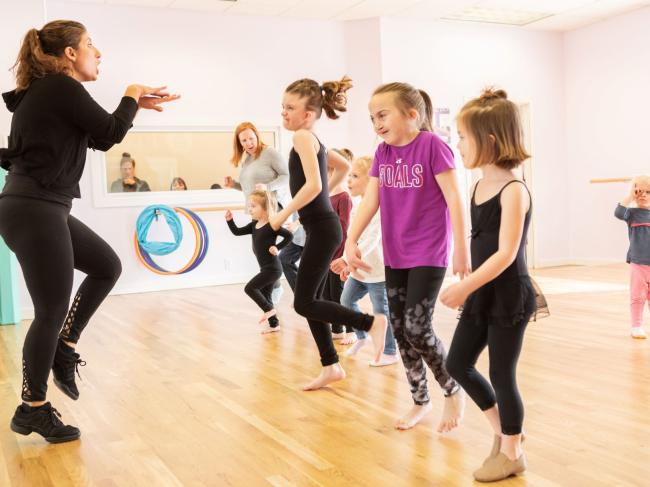Dance instructor wearing all black poses with one leg up in front of six children in a similar pose