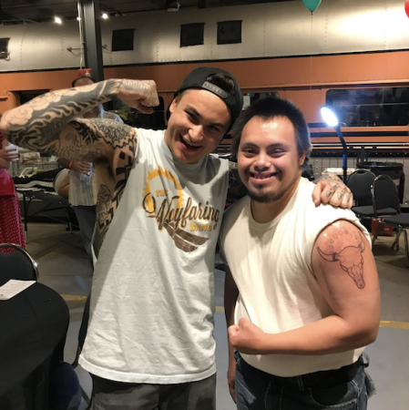 Alt text: Justin Wild (a tattoo artist who joined the tour) and Jake Blackgoat are flexing their arm muscles to show off their tattoos. Justin has a full sleeve of tattoos and Jake has a tattoo of a cow skull on his upper bicep.