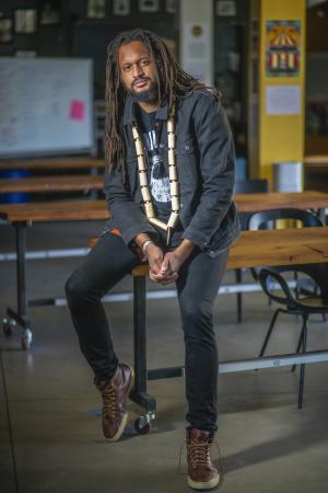 Stephen Bracket, an African American male in his 30s with dreadlocks in an all black outfit with a white large-bead necklace and brown sneakers, sits on a wooden desk in a school classroom