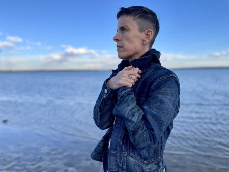 A white nonbinary person with a short haircut and jean jacket stands with left profile facing the viewer in front of a lake and partially cloudy sky