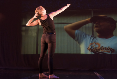 Bridget Heddens is in the foreground, wearing black and facing away from the camera. She has her left hand on the back of her head and her right arm extended. In the background, an image is projected of a person with a Wayfairing Band t-shirt and a hat. He has his hand on his head and is looking up to the sky.