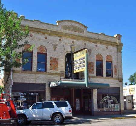 View of exterior of 1923 Rialto theater taken from across the street. The building is two stories of light colored brick A marquee with blurred writing announces a show and a white and a red car are parked out front. To the left of the cars part of a tree peers over.