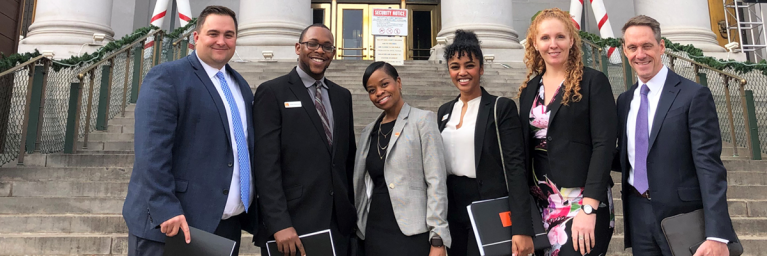 Six FirstBank employees dressed in suits or a blouse/skirt smiling and standing in front of the bottom of the stairs of Colorado's capital building