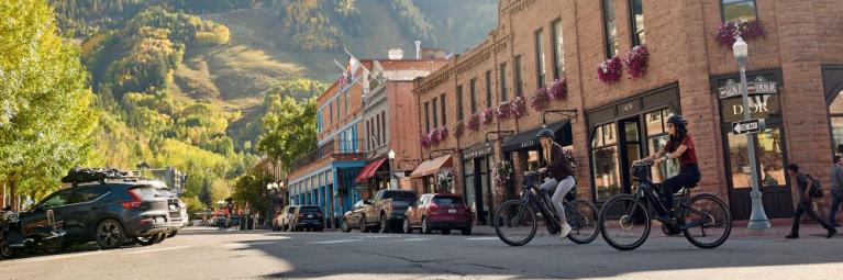 Two bicyclists ride through a downtown area during the day with mountains in the background.