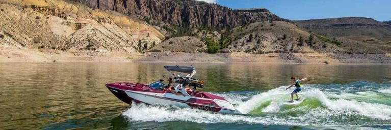 a dark red and white boat going fast on the water in a lake, with someone wakeboarding off of the back of the boat. water is splashing high behind them on this sunny clear day with a evergreen backdrop