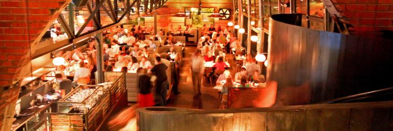 well lit restaurant with crowds of people, which is under an arch made of bricks. You can see an open concept of a kitchen and some tables 