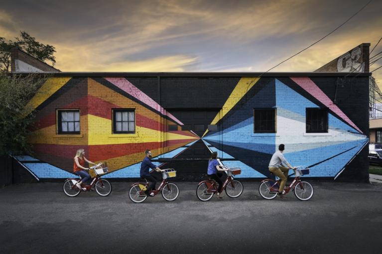 Bicyclists ride in front of a mural at sunset.