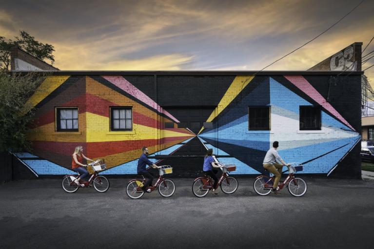 Four bicyclists ride in front of a mural of two 3D houses painted on a building, at sunset.