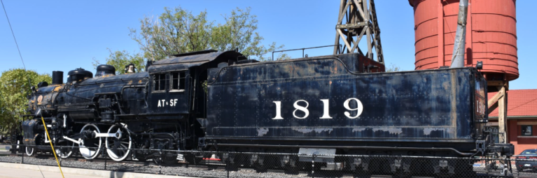 A vintage black train sits on display on tracks in front of a red silo.