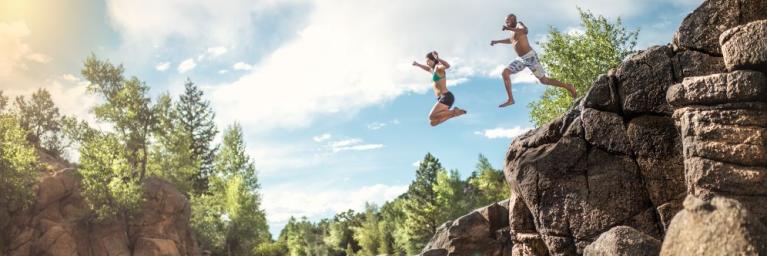 Two people jump off a rocky outcropping in to a river on a sunny day.