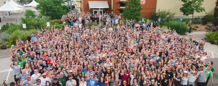 Large crowd of employees stand outside a building, looking upward and smiling