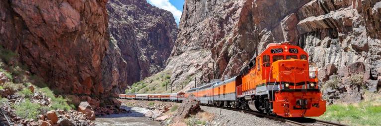 An orange train passes through a deep canyon on a sunny day.