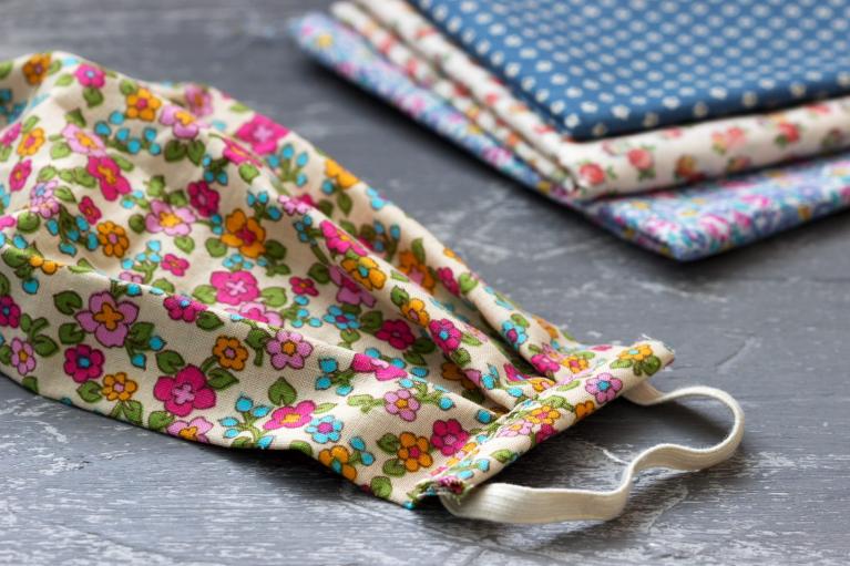 Floral print handmade mask sits on a table next to fabric that will soon be made into a mask as well.