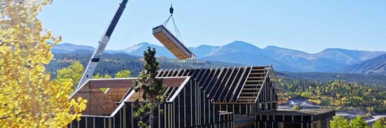 Construction of house. There is a crane dropping materials on the roof of the house. The background of the image is of a mountain range. The setting of the house is located in the green outdoors. The image is courtesy of construction firm, Phoenix Haus. 