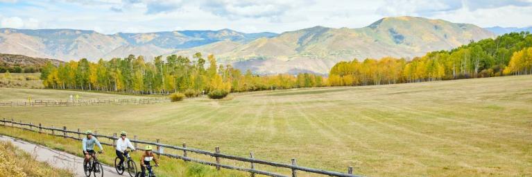 A family of four rides their bikes down a paved path in a green mountain valley.