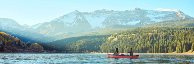 Two people canoe through a blue lake with mountains and forest in the background.