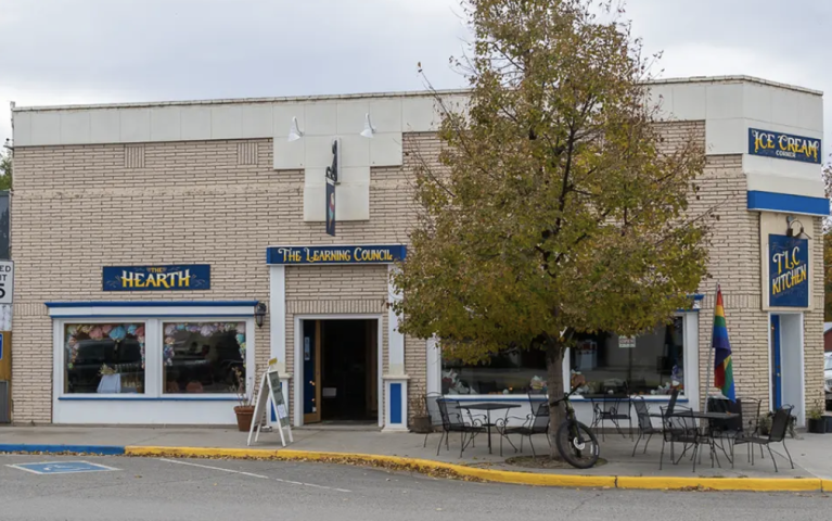 Exterior photo of tan brick building taken from across the street. Signs on the building say "the learning council kitchen" "the hearth" and "ice cream". It is a cloudy day and a leafy tree and tables, chairs, and a rainbow flag can be seen in front of the building.