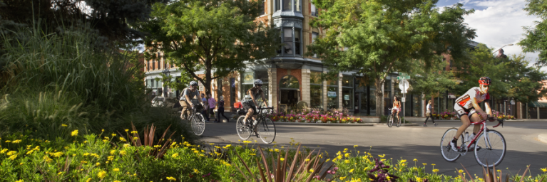 Bicyclists pass through a grassy and flower filled part of Downtown Ft. Collins, with a brown building the the background