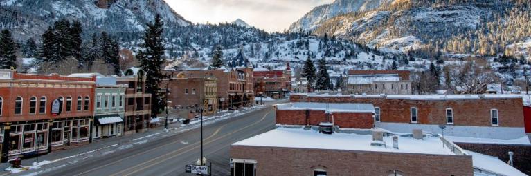 Downtown Ouray with buildings on either side covered in a light dust of snow, snowy mountain tops in the background and sun shining through onto the street